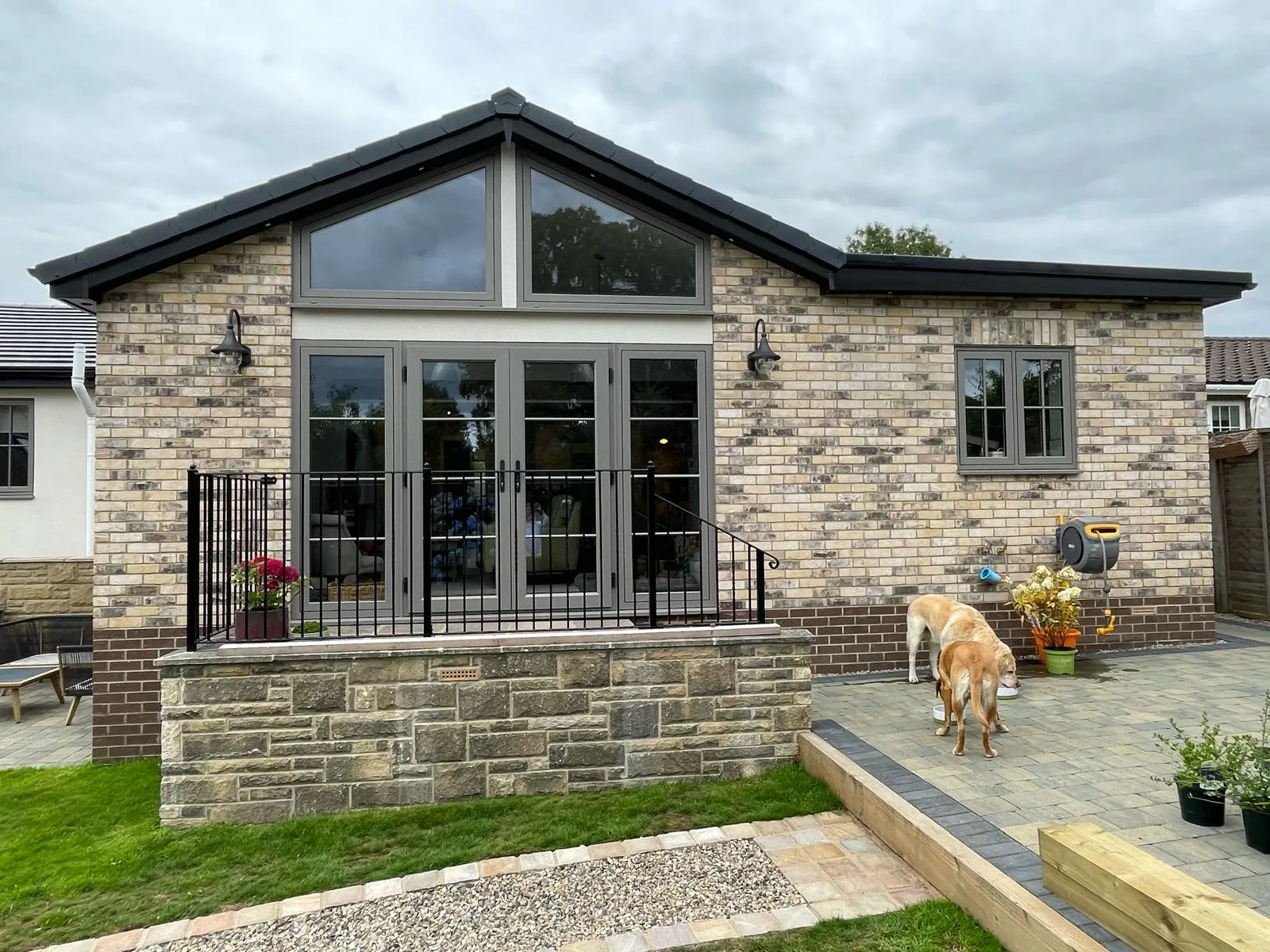 A modern brick house with a gabled roof, large glass doors, and a fenced patio. Two dogs are exploring the yard.